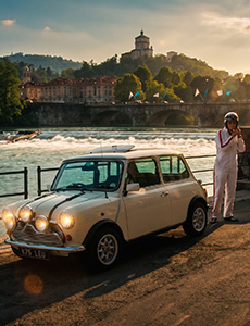 Una Mini Cooper d'epoca color crema accanto alla diga sul Po a Torino nella luce del tramonto, con la chiesa della Gran Madre di Dio sulla collina alle spalle